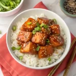 A bowl of crispy honey garlic salmon bites served over fluffy jasmine rice, garnished with sesame seeds and fresh chives, with chopsticks on the side.